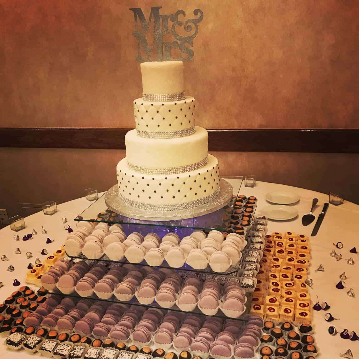 A four-tier white wedding cake with “Mr & Mrs” topper, surrounded by trays of purple macarons and various small desserts on a round table.