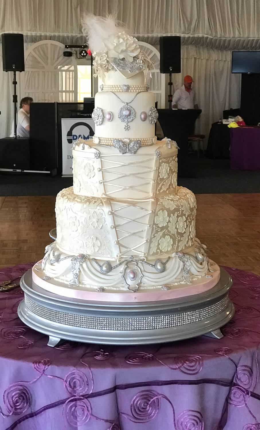 A five-tier white wedding cake with ornate lace-like decorations, pearls, and silver embellishments sits on a round table covered in a purple tablecloth.