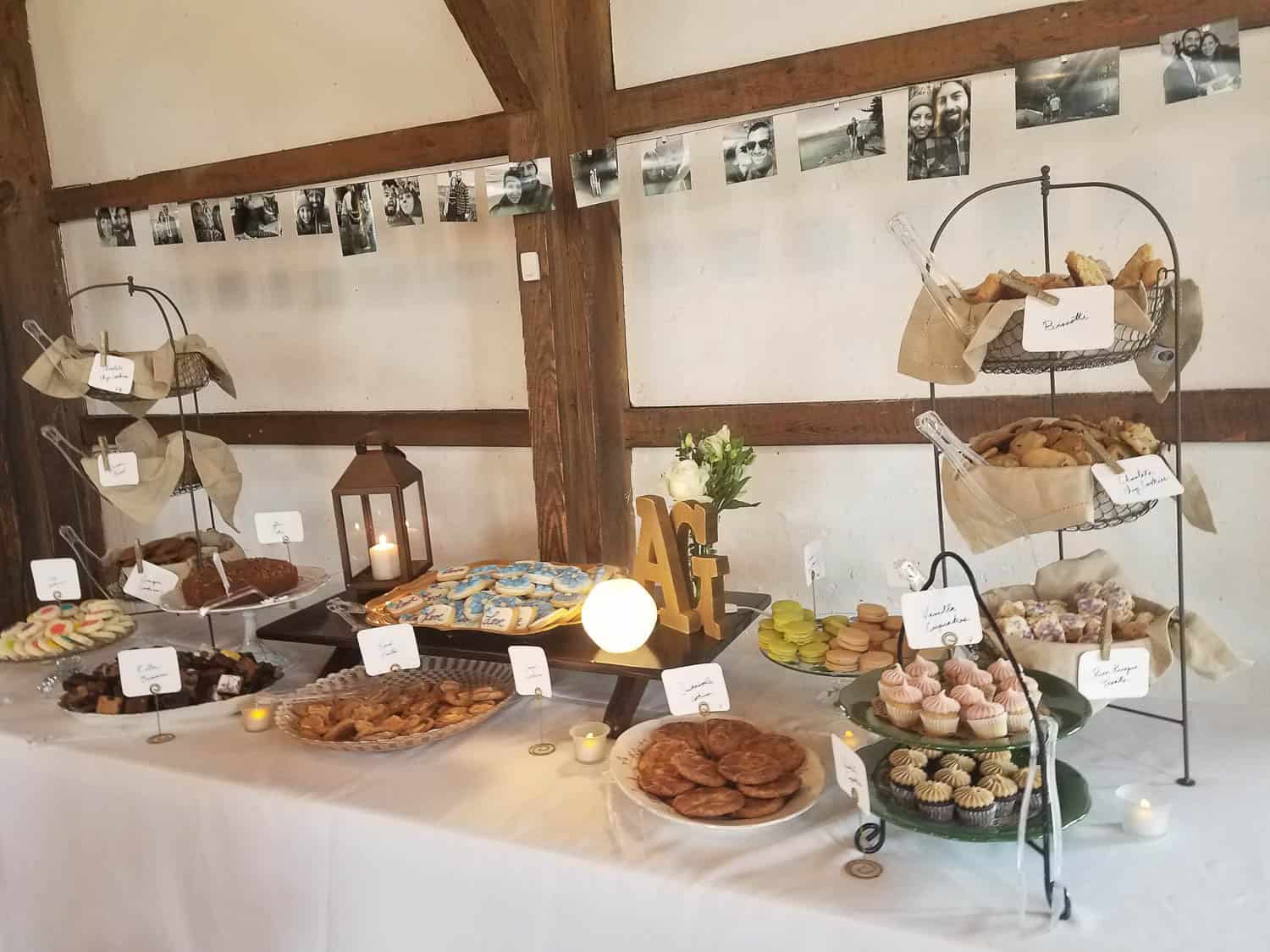 A dessert table with assorted cookies, cupcakes, and pastries displayed on tiered stands, with labeled cards, candles, and framed photos hung on the wall behind.