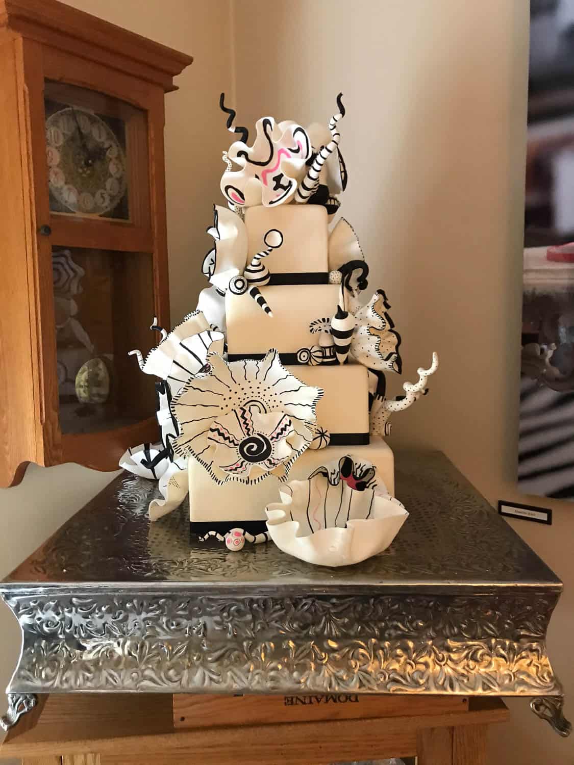 A multi-tiered square cake with abstract black and white decorations, set on a silver stand next to a wooden cabinet and a white ceramic bowl.