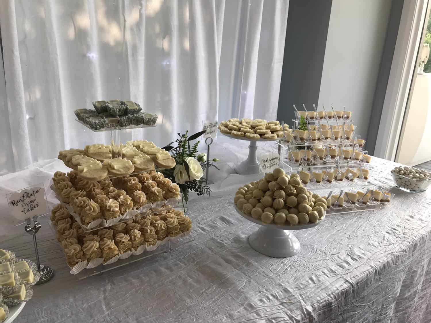 A dessert table displays an assortment of pastries, cupcakes, cake pops, and truffles on stands, with a white tablecloth and floral decorations.