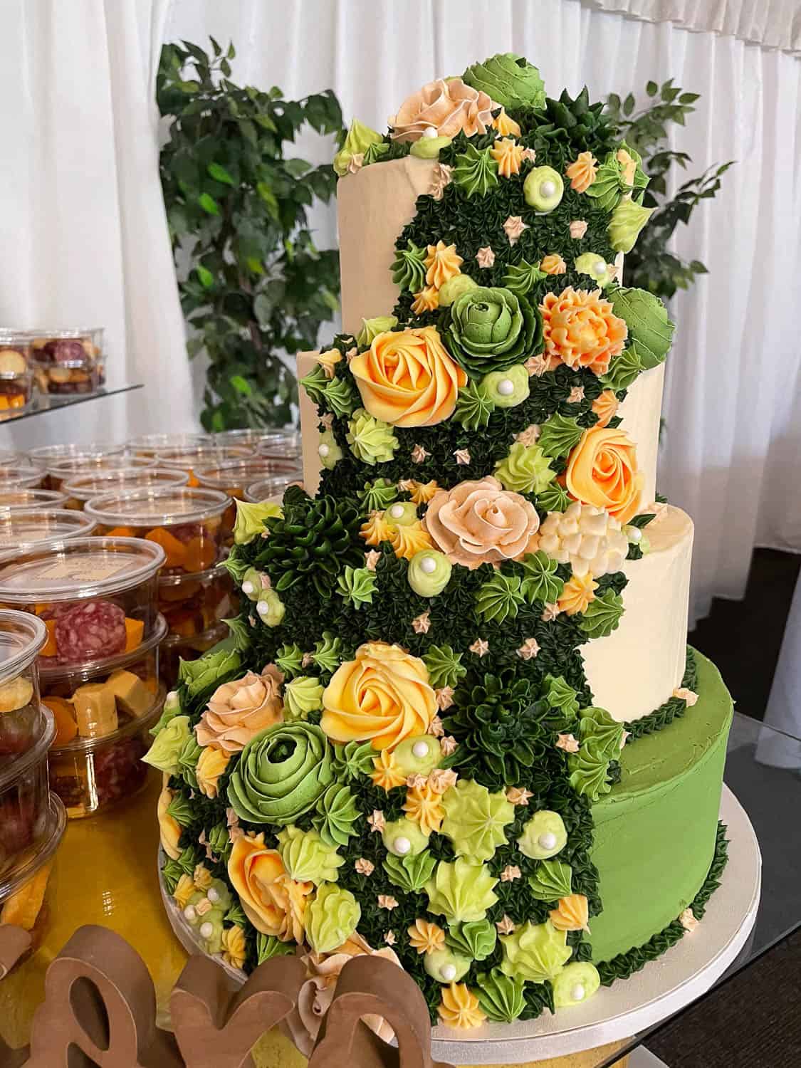 Three-tiered cake decorated with cascading buttercream flowers in yellow, peach, and green, surrounded by greenery and pearls, and fruit cups in the background.