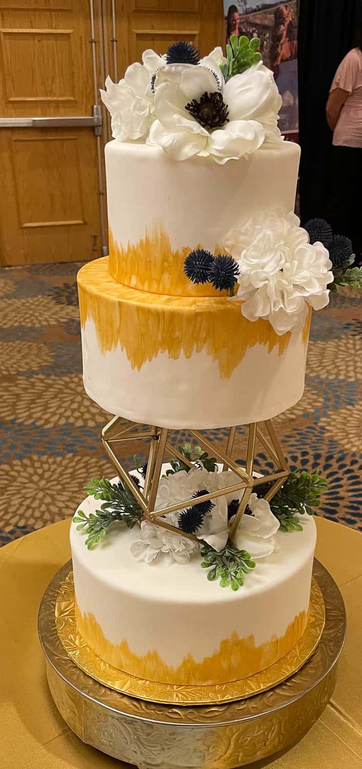 3-tiered white cake with gold brushstroke accents, decorated with white and dark blue flowers, and a geometric gold stand between the middle and bottom tiers.