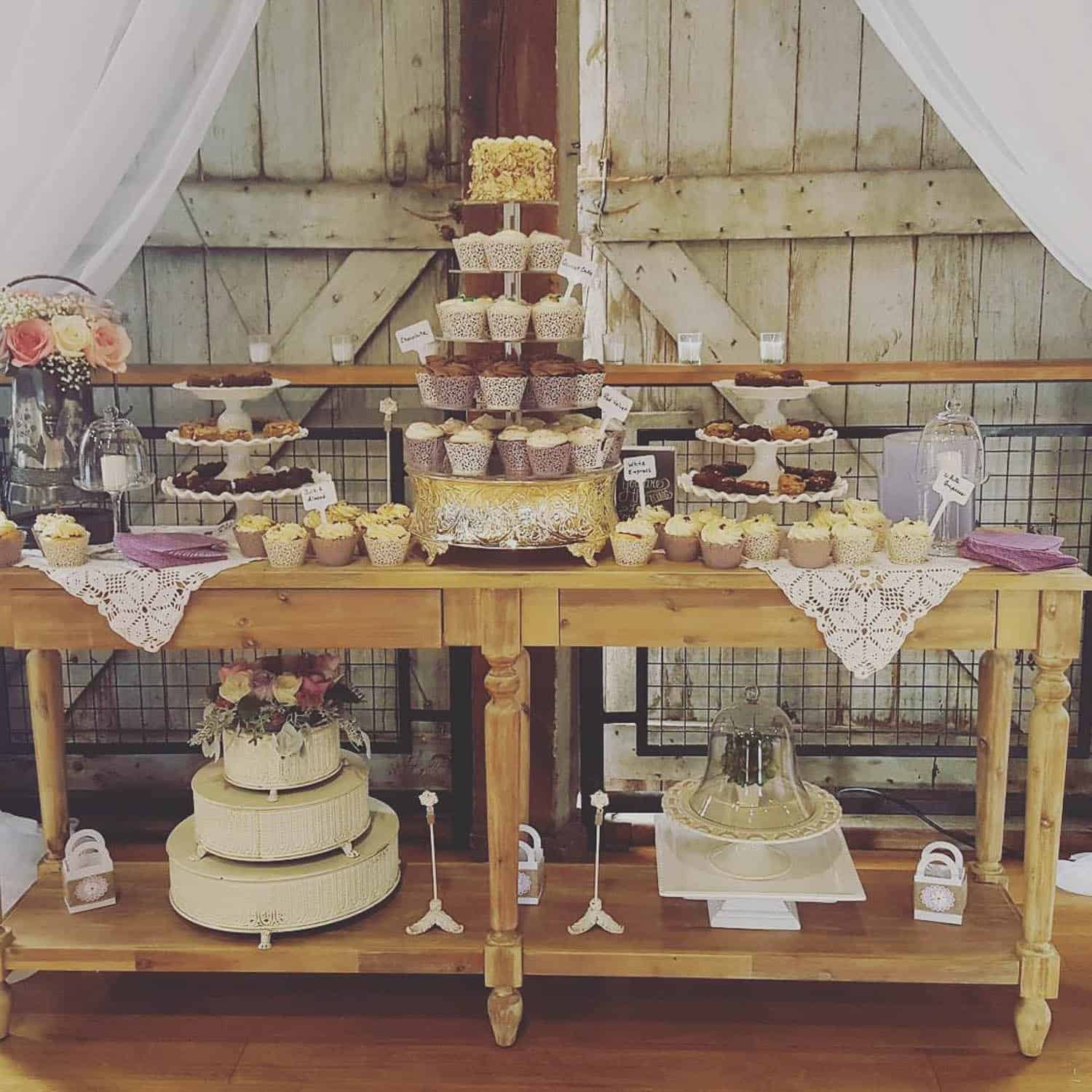 A dessert table features a cupcake tower, trays of cupcakes, a decorated cake, lace doilies, and floral arrangements against a rustic wooden barn backdrop.