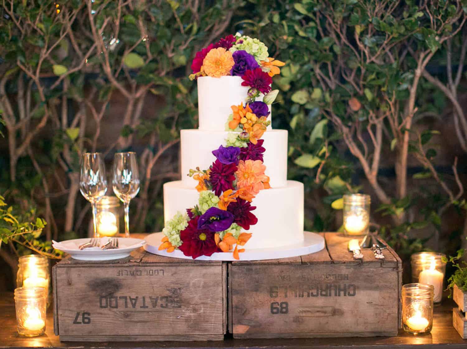 Three-tier white cake decorated with vibrant flowers, displayed on wooden crates, surrounded by candles and glassware, with greenery in the background.