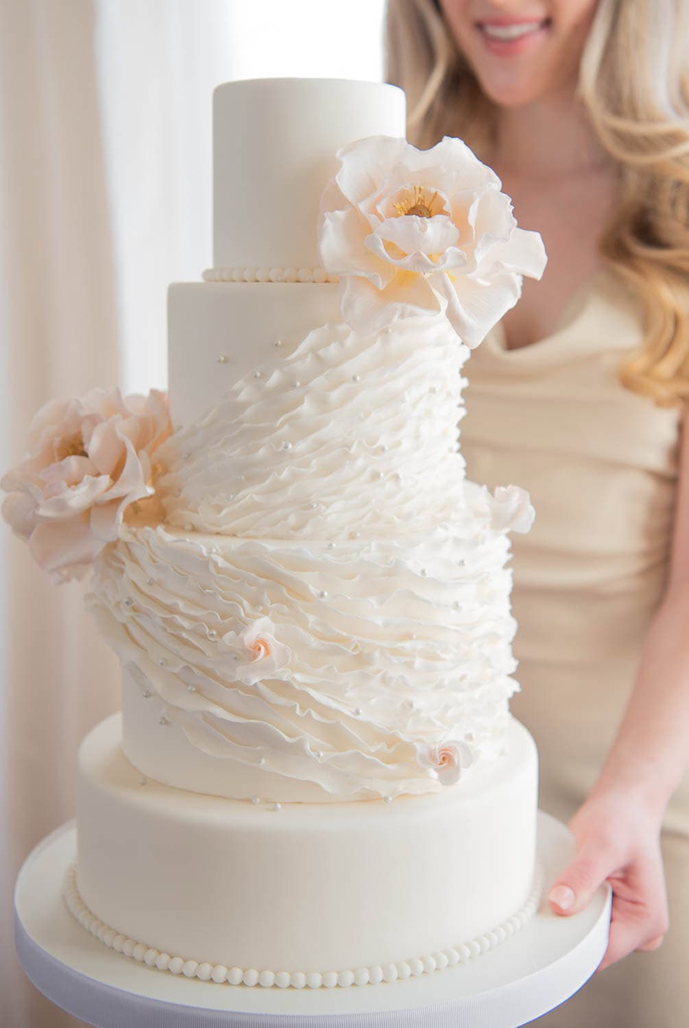 A three-tiered white wedding cake with ruffled icing and large pale flowers, held by a woman in a cream dress.