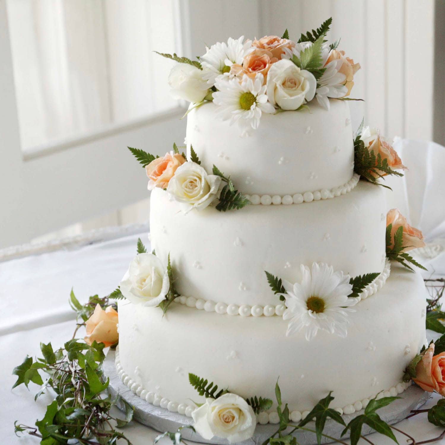 Three-tiered white cake decorated with white and peach flowers, green leaves, and piped pearl-like details, displayed on a white tablecloth.