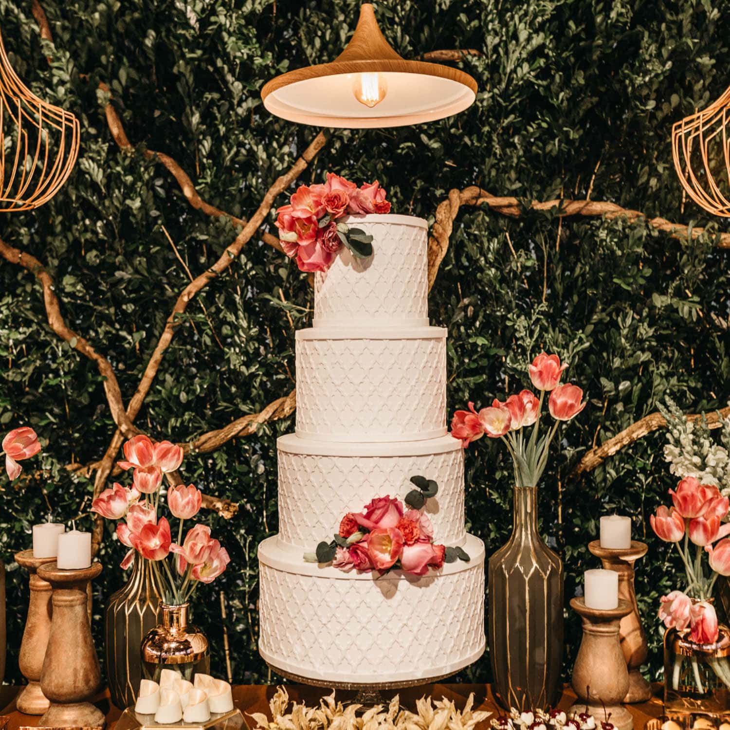 Three-tiered white cake with quilted pattern, pink flowers, and greenery, surrounded by candles and vases on a wooden table with green foliage backdrop.