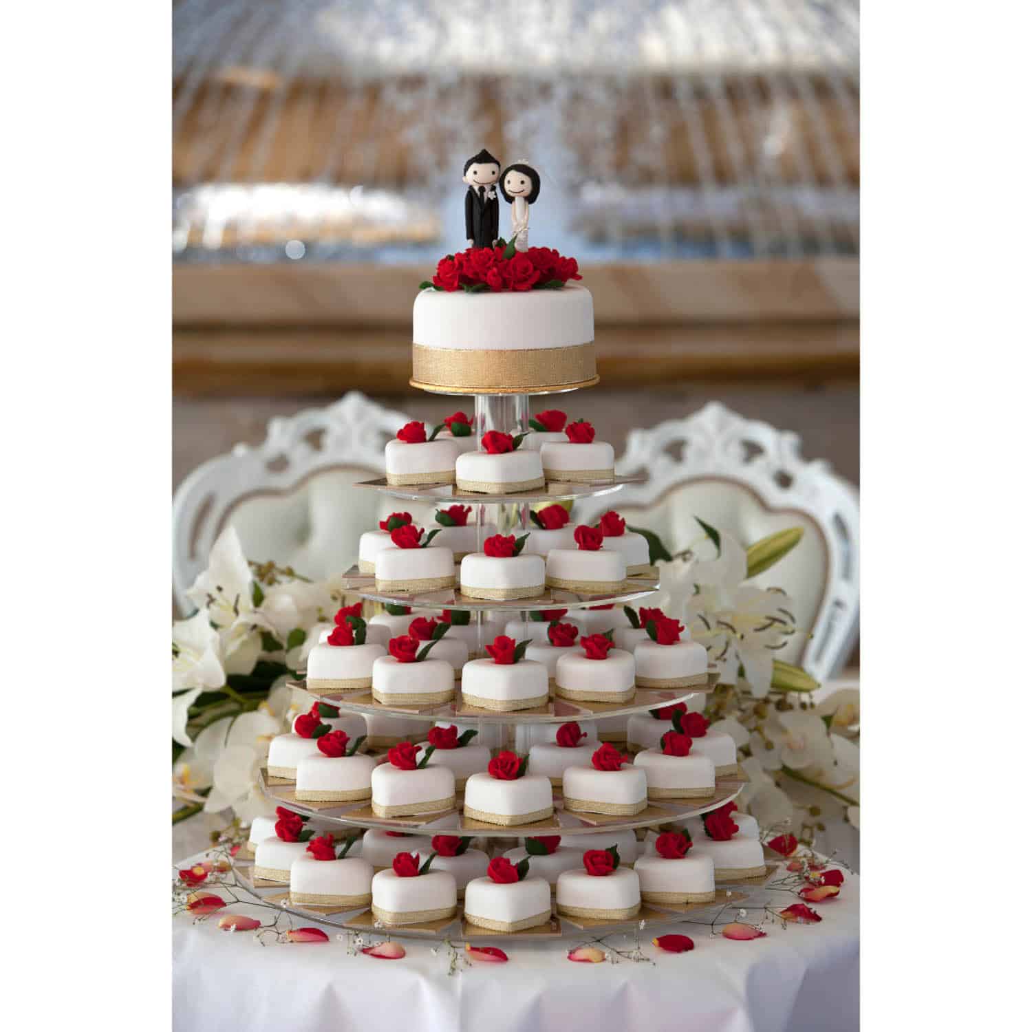 A tiered wedding cake display with white and red mini cakes, topped with a small cake featuring bride and groom figurines and red flowers.