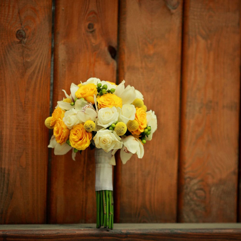 A bouquet of yellow and white flowers with green stems is wrapped in white ribbon and placed in front of a wooden background.