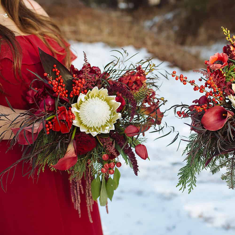 A person in a red dress holding a large bouquet with red flowers, greenery, berries, and a white protea; snow-covered ground in the background.