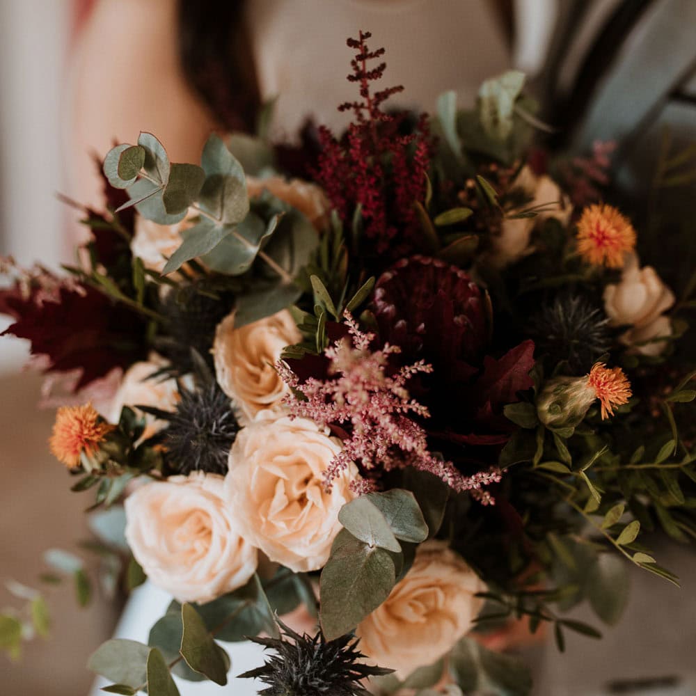 A close-up of a bouquet featuring cream roses, dark red flowers, orange thistles, and various green foliage.