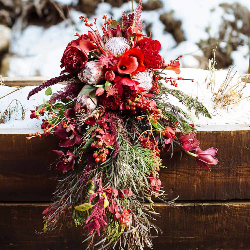 A cascading bouquet of red and pink flowers with greenery and berries rests on a wooden ledge dusted with snow.
