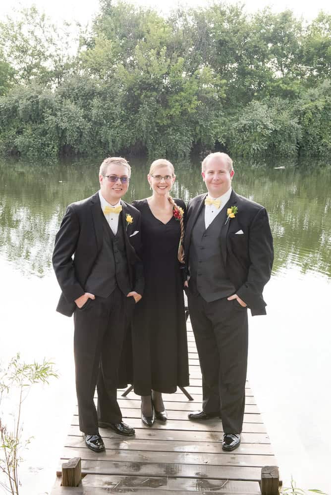 Three people dressed formally stand on a wood dock by a river, with trees in the background. Two men wear tuxes and a woman wears a long black dress and glasses.