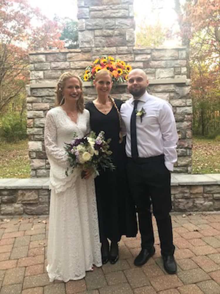 Three people pose in front of a stone wall outdoors; one in a white wedding dress holding a bouquet, one in a black dress, and one in a white shirt with a tie.