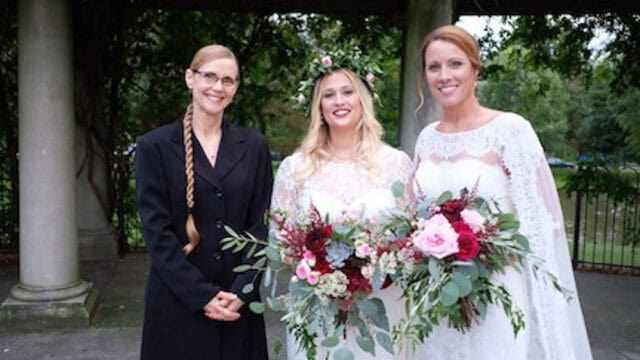 Three women stand outdoors; two in white dresses holding bouquets, and one in a black outfit. The background features stone columns and greenery.
