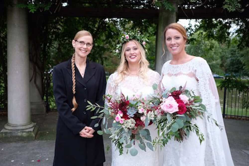 Three women stand outdoors; two in white dresses holding bouquets, and one in a black outfit. The background features stone columns and greenery.