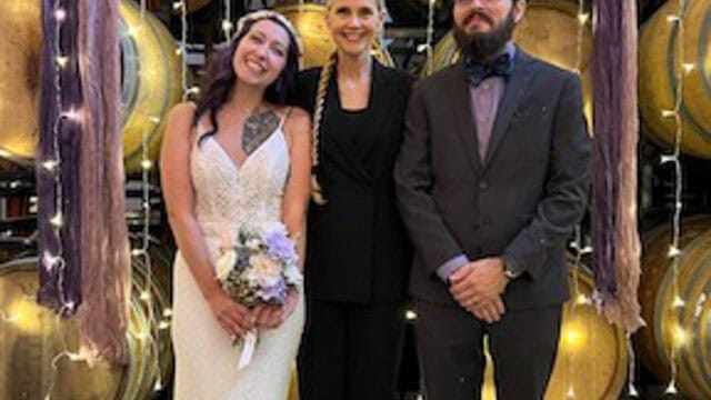 Three people stand before stacked wine barrels: a woman in a wedding dress, a woman in a black suit, and a man in a dark suit.