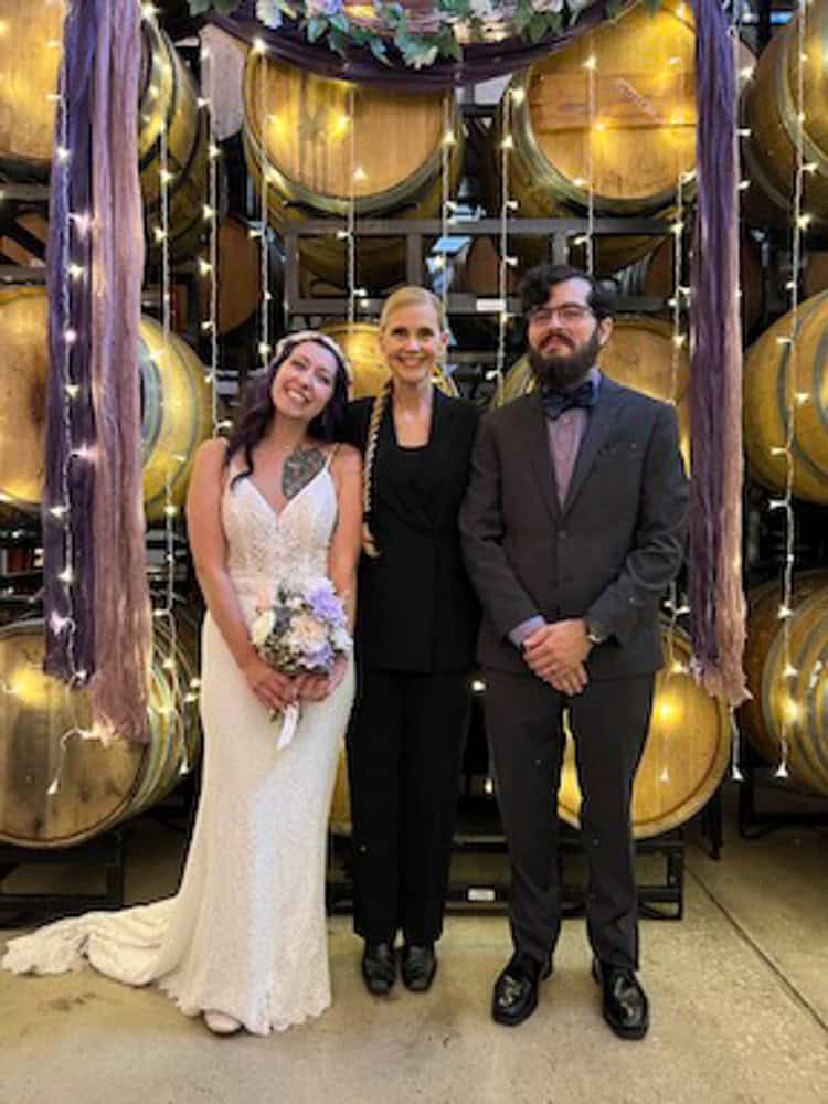 Three people stand before stacked wine barrels: a woman in a wedding dress, a woman in a black suit, and a man in a dark suit.