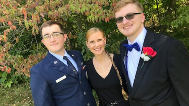 Three people pose outdoors: one in a military uniform, one in a suit with a red boutonniere, and one in a black dress, surrounded by trees and greenery.