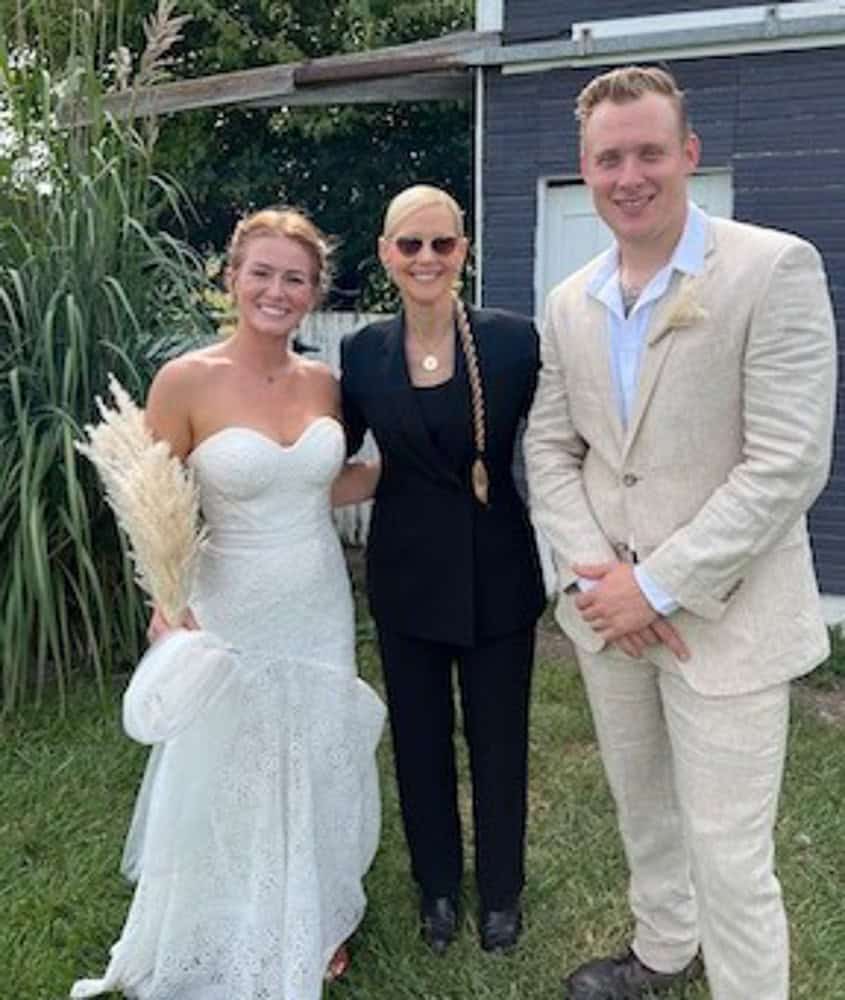 A bride in a white dress, a woman in a black suit, and a groom in a beige suit stand together outdoors on grass, smiling at the camera.