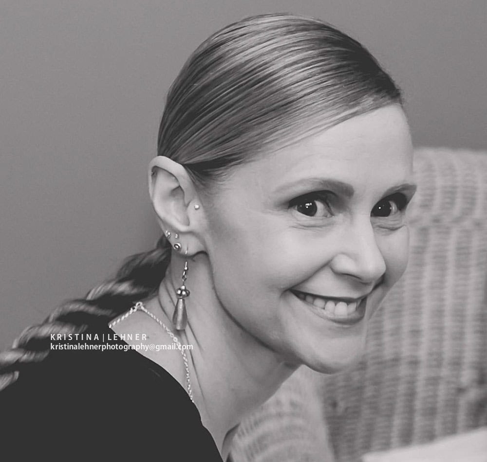 A woman with light hair pulled back in a braid smiles at the camera; she is wearing earrings and sitting in front of a wicker chair.