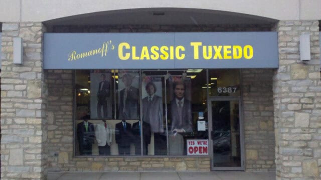 Storefront of Romanoff’s Classic Tuxedo with large window displays featuring mannequins in tuxedos and a sign that reads “YES WE’RE OPEN.”.