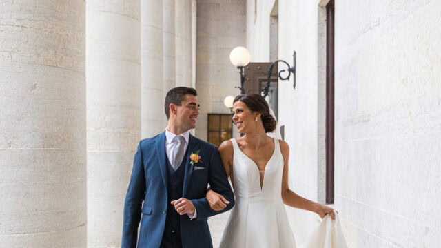 A bride in a white gown and a groom in a blue suit walk arm in arm down a marble corridor with large columns, smiling at each other.