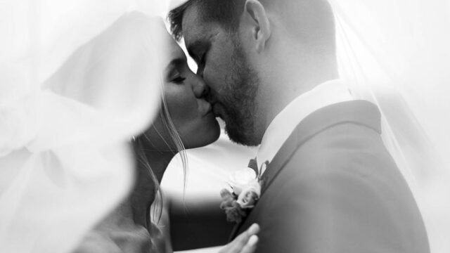 A bride and groom kiss under a veil, with soft light and blurred edges creating a romantic, intimate scene.