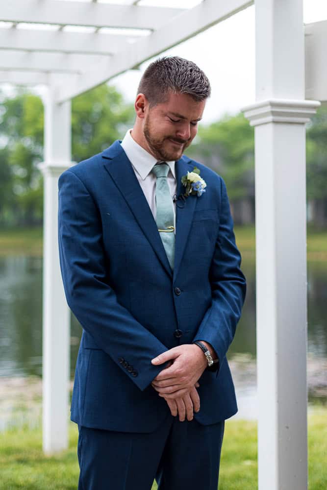 A man in a blue suit stands outdoors under a white pergola, looking down with his hands clasped in front of him.