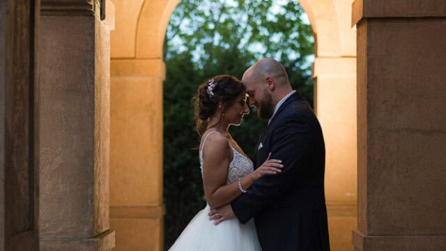 A bride and groom stand close together under an archway, illuminated by a hanging lantern, with greenery visible in the background.