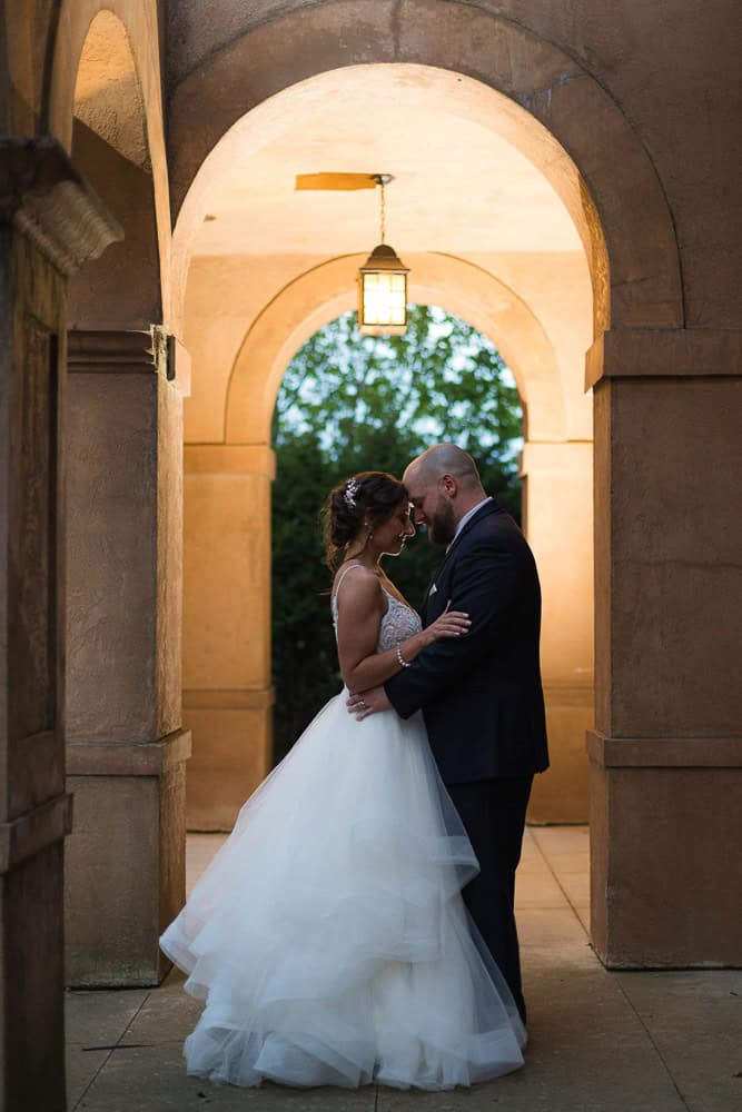 A bride and groom stand close together under an archway, illuminated by a hanging lantern, with greenery visible in the background.