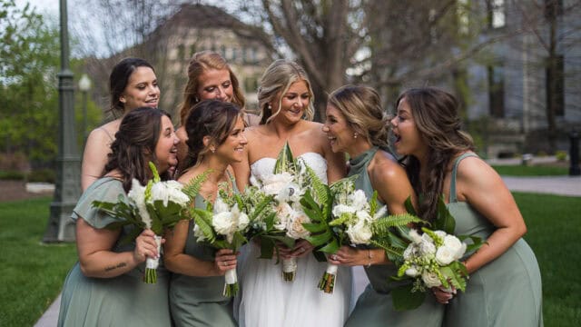 Seven women in formal dresses, six in sage green and a bride in white, stand outdoors holding floral bouquets and smiling together.