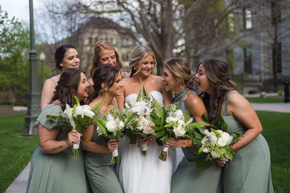 Seven women in formal dresses, six in sage green and a bride in white, stand outdoors holding floral bouquets and smiling together.
