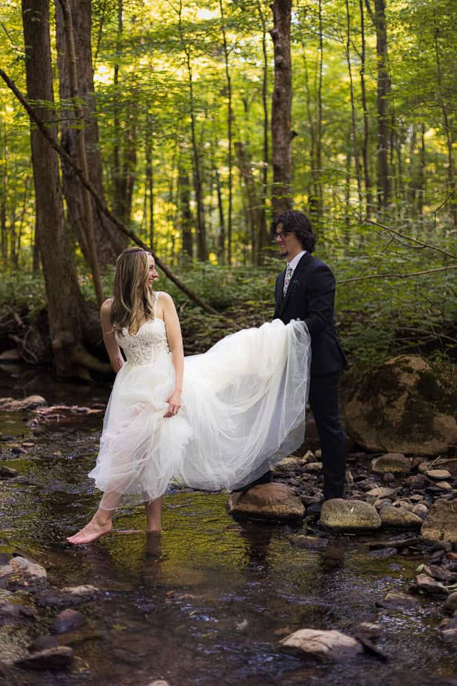 A bride stands barefoot in a shallow forest stream while a groom in a suit holds up her dress. Both are smiling and surrounded by trees.
