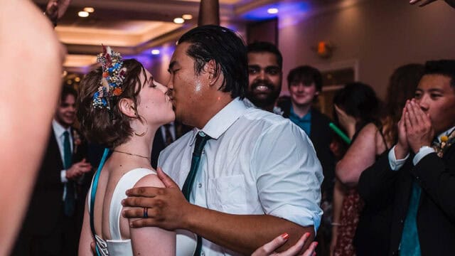 A bride and groom kiss on the dance floor at their wedding reception, surrounded by cheering and smiling guests.