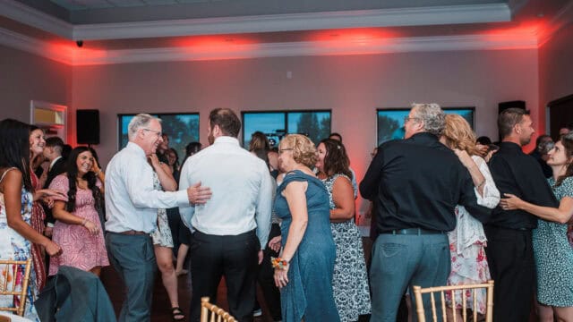 A group of people, some smiling and talking, gather closely together on a dance floor in a decorated indoor event space.
