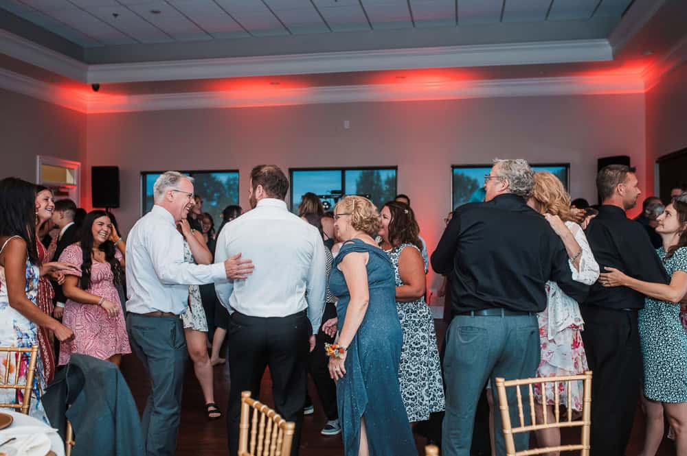 A group of people, some smiling and talking, gather closely together on a dance floor in a decorated indoor event space.