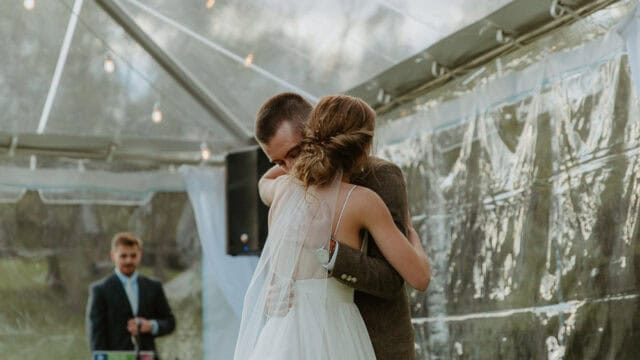 A bride and groom embrace during their wedding reception under a clear tent, with a man in a suit standing in the background near audio equipment.