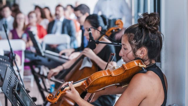 A woman plays the violin in a seated ensemble, with other musicians and an audience visible in the background inside a bright room.