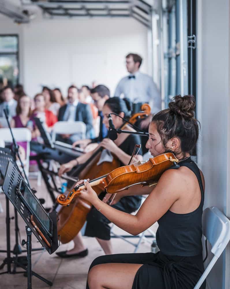 A woman plays the violin in a seated ensemble, with other musicians and an audience visible in the background inside a bright room.