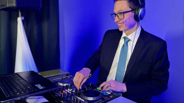 A man in a suit and tie wearing headphones is DJing with a mixer and laptop in a room with blue lighting.