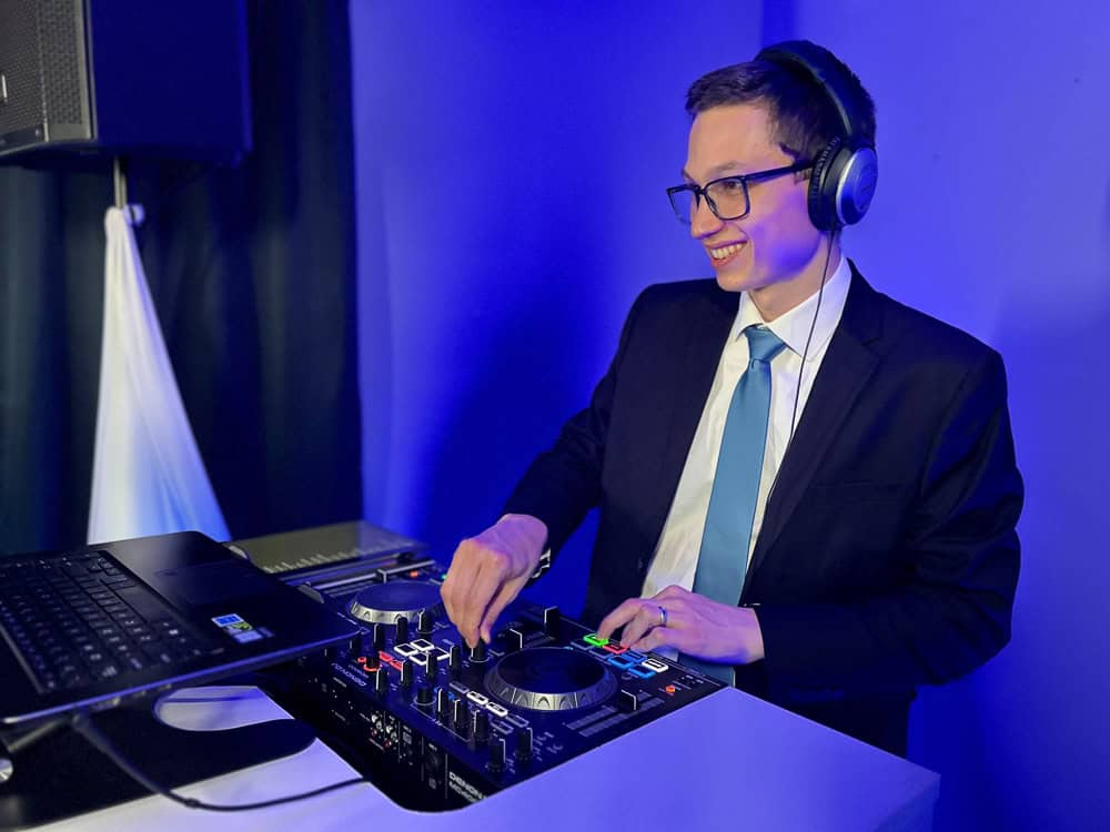A man in a suit and tie wearing headphones is DJing with a mixer and laptop in a room with blue lighting.