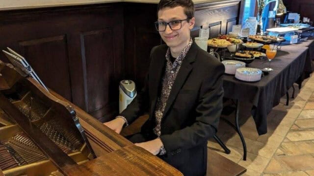 A man in a suit sits at a grand piano, smiling at the camera, with a buffet table and windows in the background.