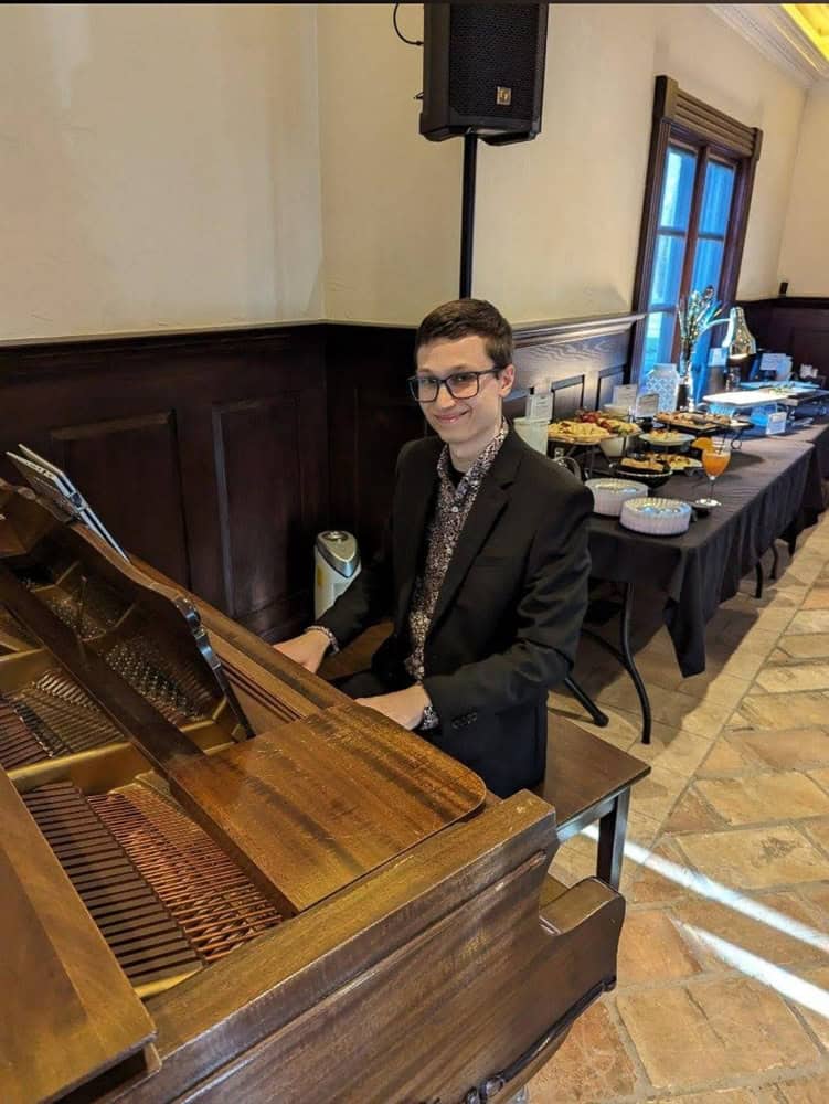 A man in a suit sits at a grand piano, smiling at the camera, with a buffet table and windows in the background.