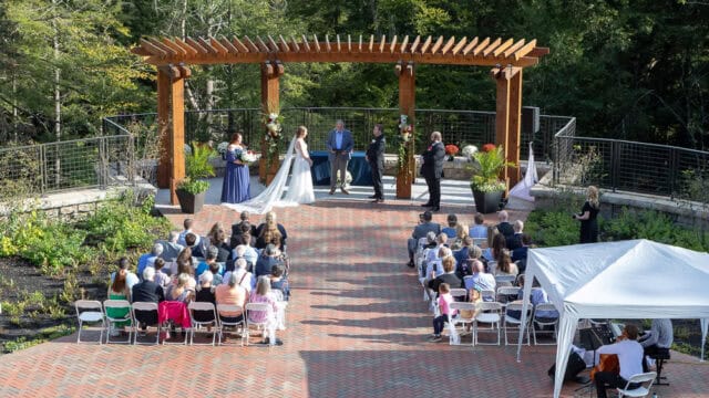 Outdoor wedding ceremony with guests seated on white chairs facing a wooden pergola where the couple and officiant stand, surrounded by greenery.