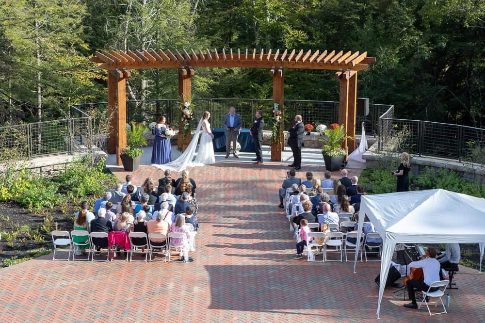 Outdoor wedding ceremony with guests seated on white chairs facing a wooden pergola where the couple and officiant stand, surrounded by greenery.