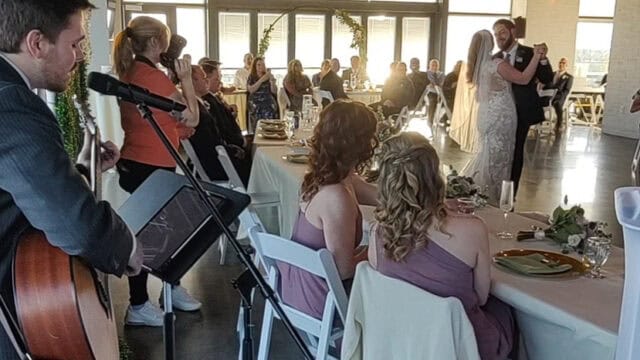 A bride and groom share their first dance in a sunlit reception hall while guests watch and a musician plays guitar and sings nearby.