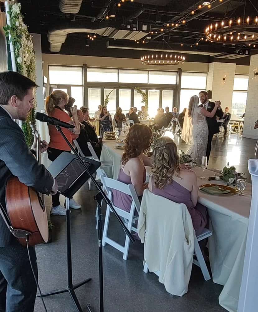 A bride and groom share their first dance in a sunlit reception hall while guests watch and a musician plays guitar and sings nearby.