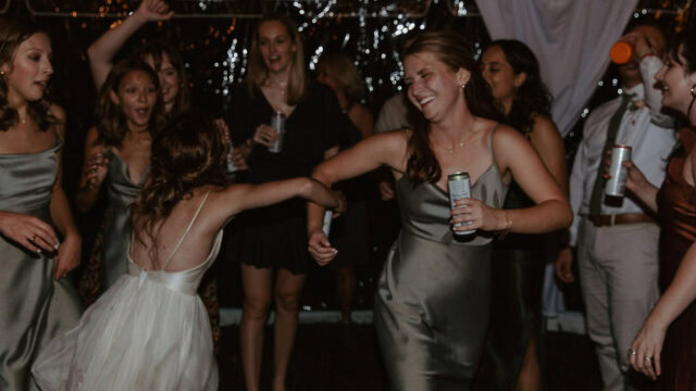 A group of people in formal attire dancing and holding drinks at a celebration, with two women in the foreground holding hands and smiling.